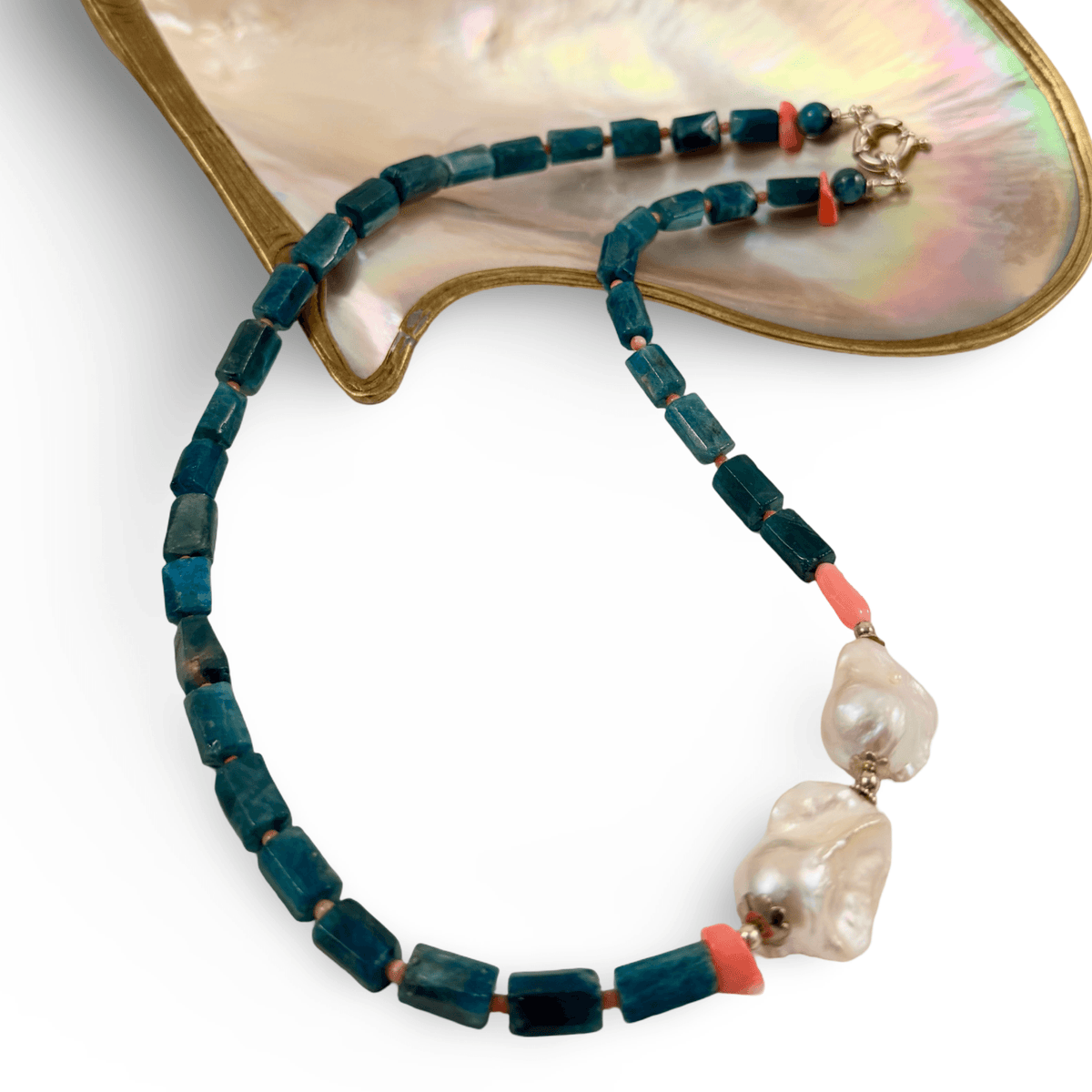A beaded necklace with dark teal rectangular beads, coral pink spacer beads, and two large baroque white pearls, displayed on a gold-tone and mother-of-pearl stand against a white background.