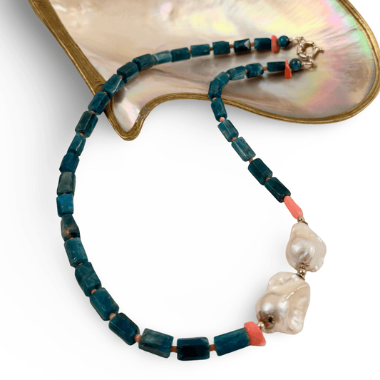 A beaded necklace with dark teal rectangular beads, coral pink spacer beads, and two large baroque white pearls, displayed on a gold-tone and mother-of-pearl stand against a white background.