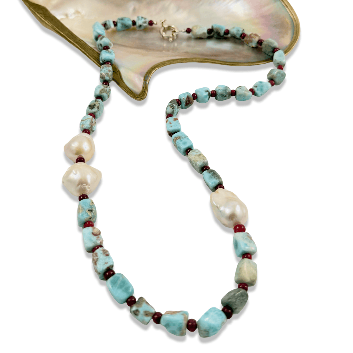A beaded necklace featuring light blue turquoise-like beads, irregular white baroque pearls, and small dark red spacer beads, displayed on a white background with a gold and mother-of-pearl shell prop.