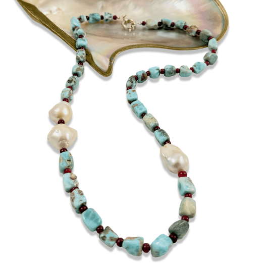 A beaded necklace featuring light blue turquoise-like beads, irregular white baroque pearls, and small dark red spacer beads, displayed on a white background with a gold and mother-of-pearl shell prop.