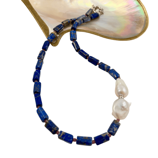 A beaded necklace featuring dark blue lapis lazuli cube beads and two baroque-style white freshwater pearls, displayed on a gold-tone and iridescent mother-of-pearl clam shell against a white background.