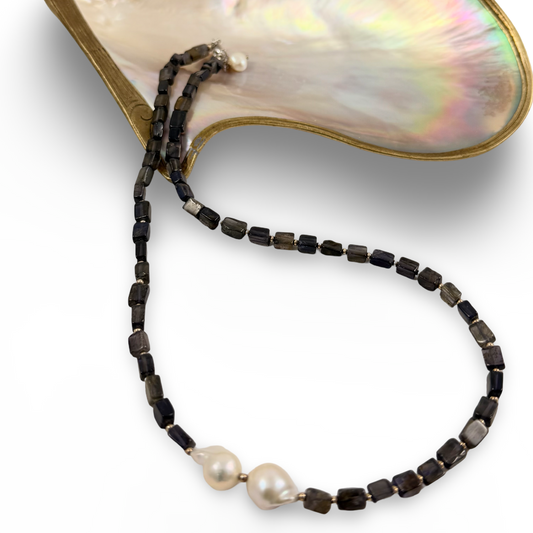 A beaded necklace with dark faceted beads, two white baroque pearls, and a gold-tone clasp, displayed on a mother-of-pearl shell prop against a white background.
