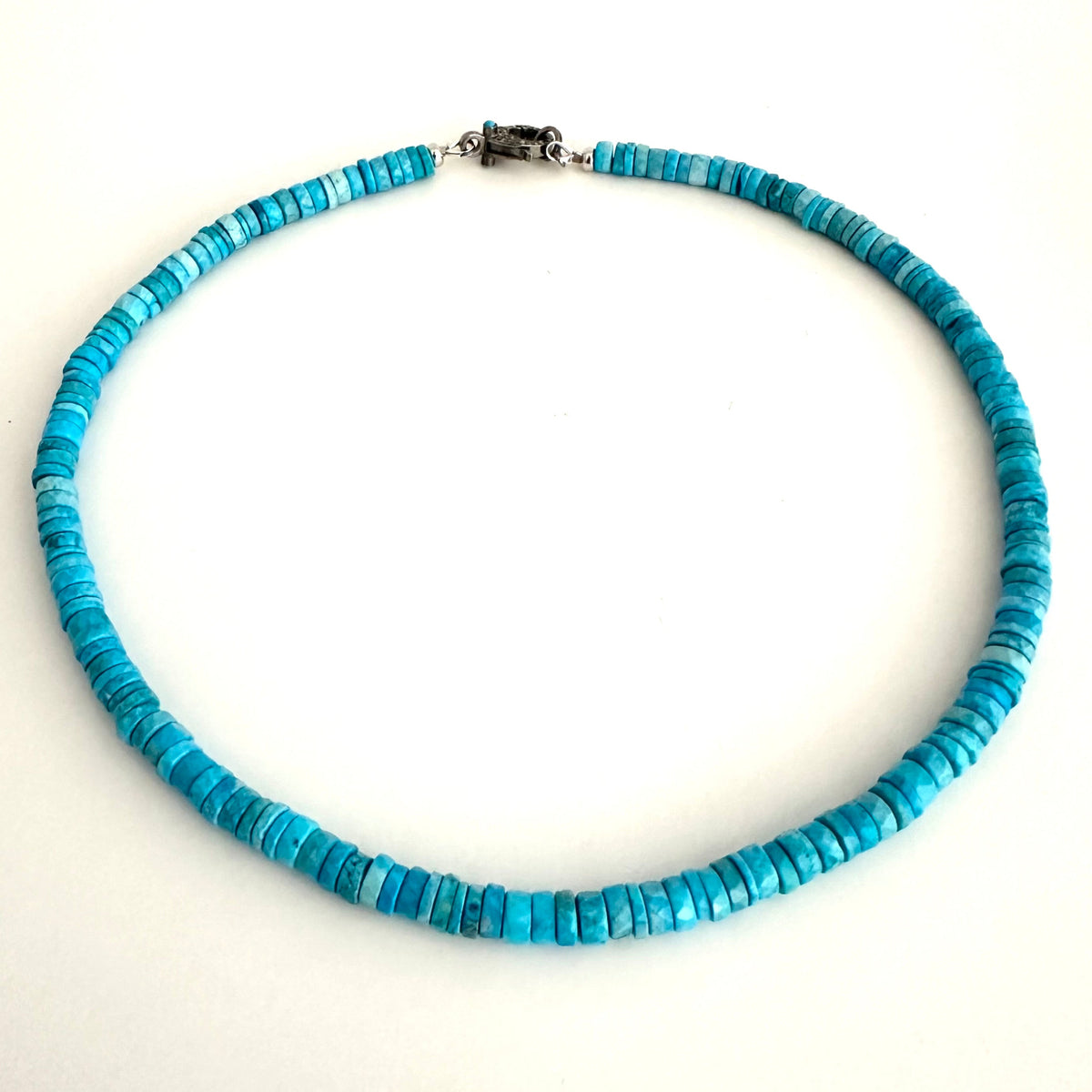 A turquoise beaded necklace made of small, flat rectangular disc-like beads, arranged in a circle on a plain white background. It features a silver-toned metal clasp at the top.