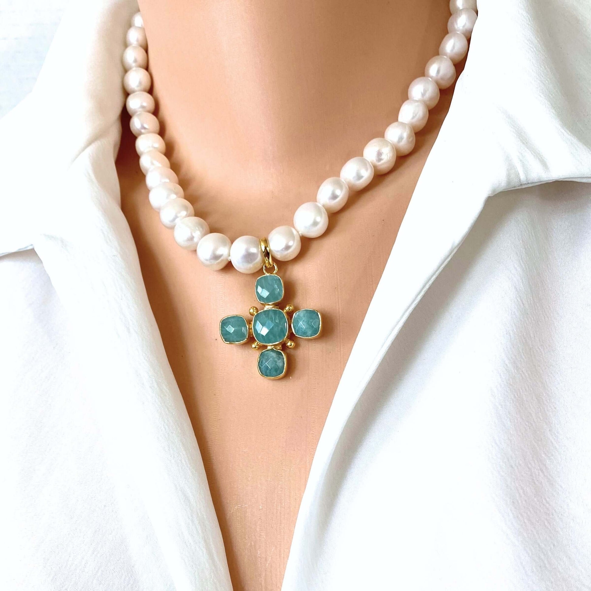 A close-up of a pearl necklace with a gold cross pendant featuring turquoise-blue stones, worn on a model's neck over a white collared shirt.