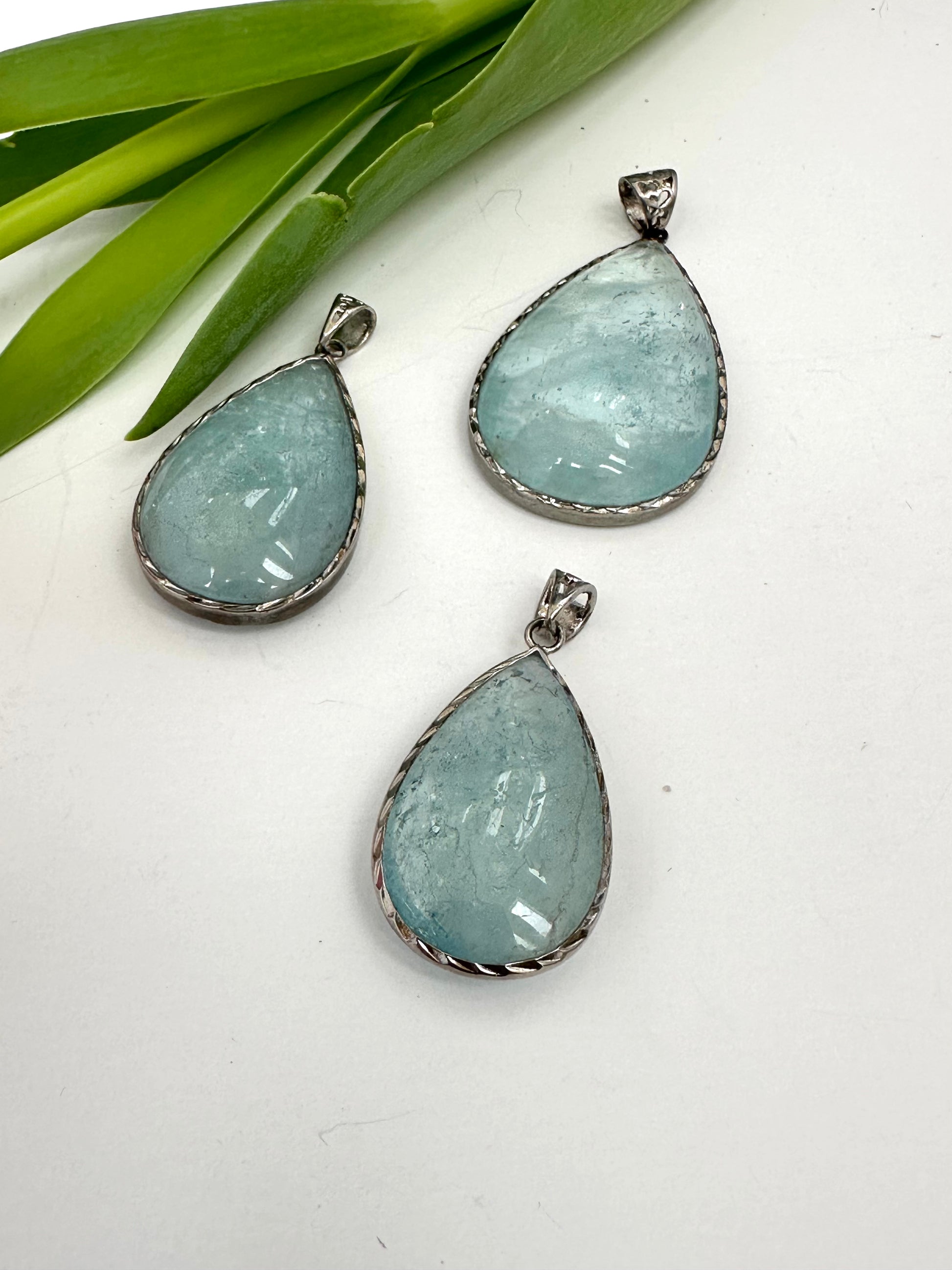 Three teardrop-shaped aquamarine pendants on a white surface with green leaves in the background.