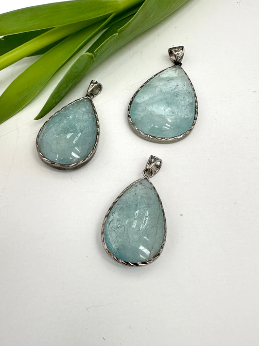 Three teardrop-shaped aquamarine pendants on a white surface with green leaves in the background.