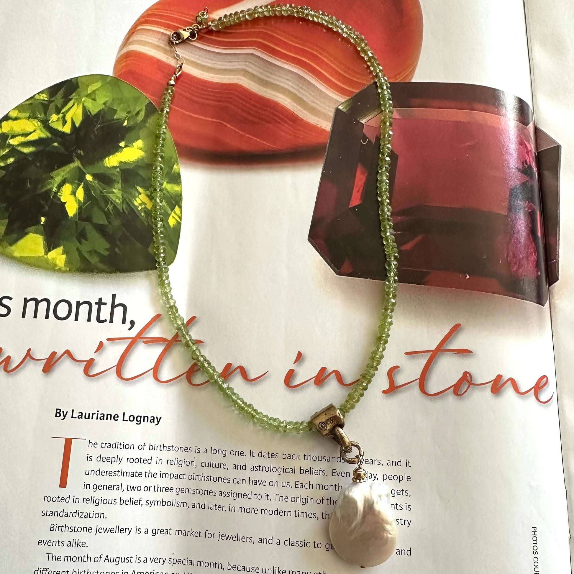 A beaded necklace with a white pearl-like pendant and green beads draped over an open magazine page featuring gemstone imagery and the text 'written in stone'.