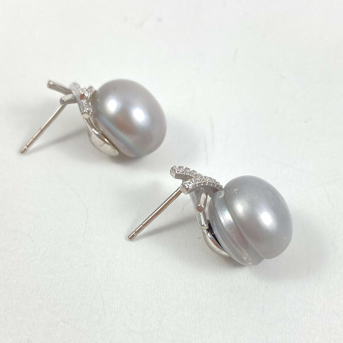 A pair of silver stud earrings featuring oval gray pearls set with small clear stones, shown on a light textured background.