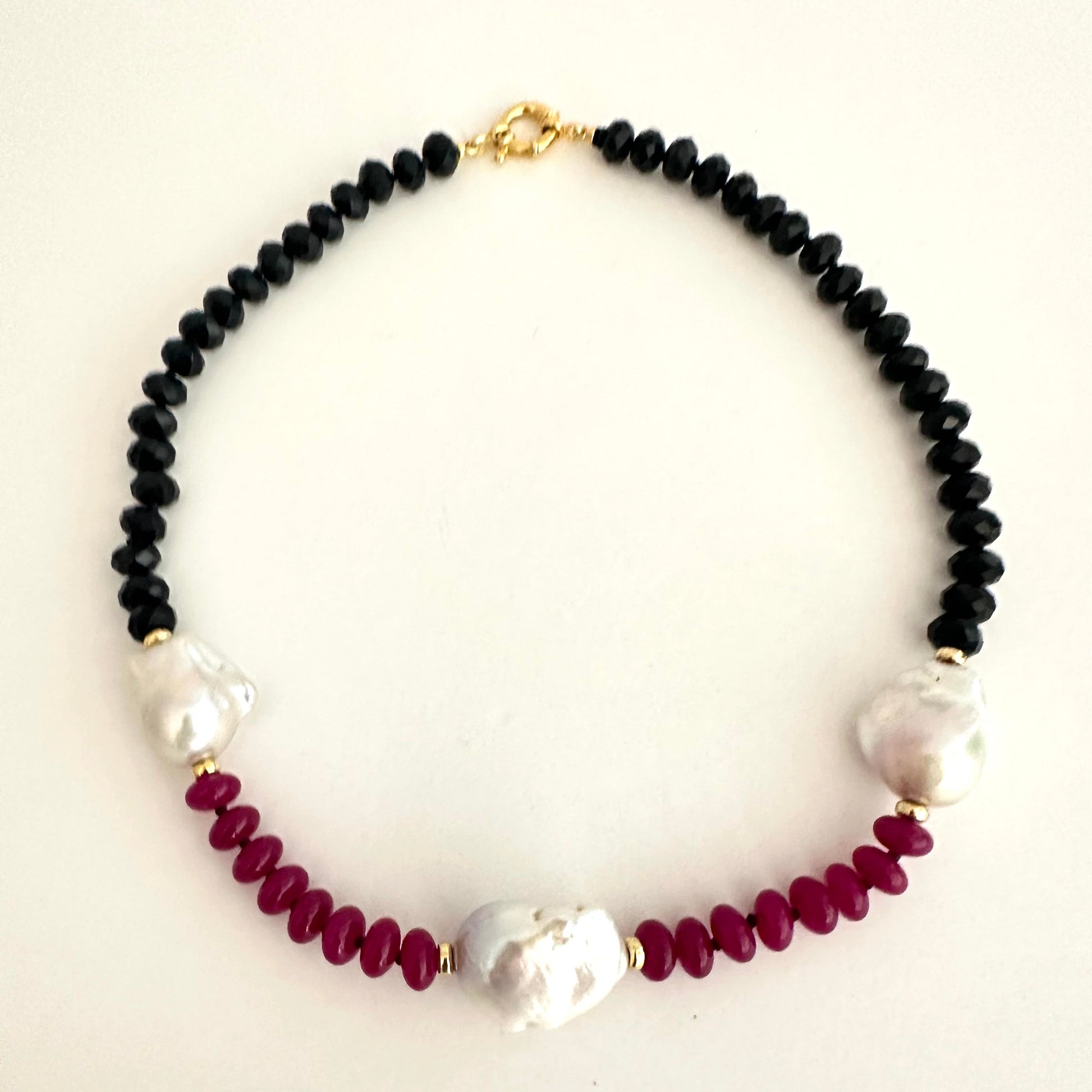 A beaded necklace featuring black faceted beads, deep burgundy round beads, and three white baroque pearls, accented with small gold spacers and a gold clasp, displayed on a light cream background.