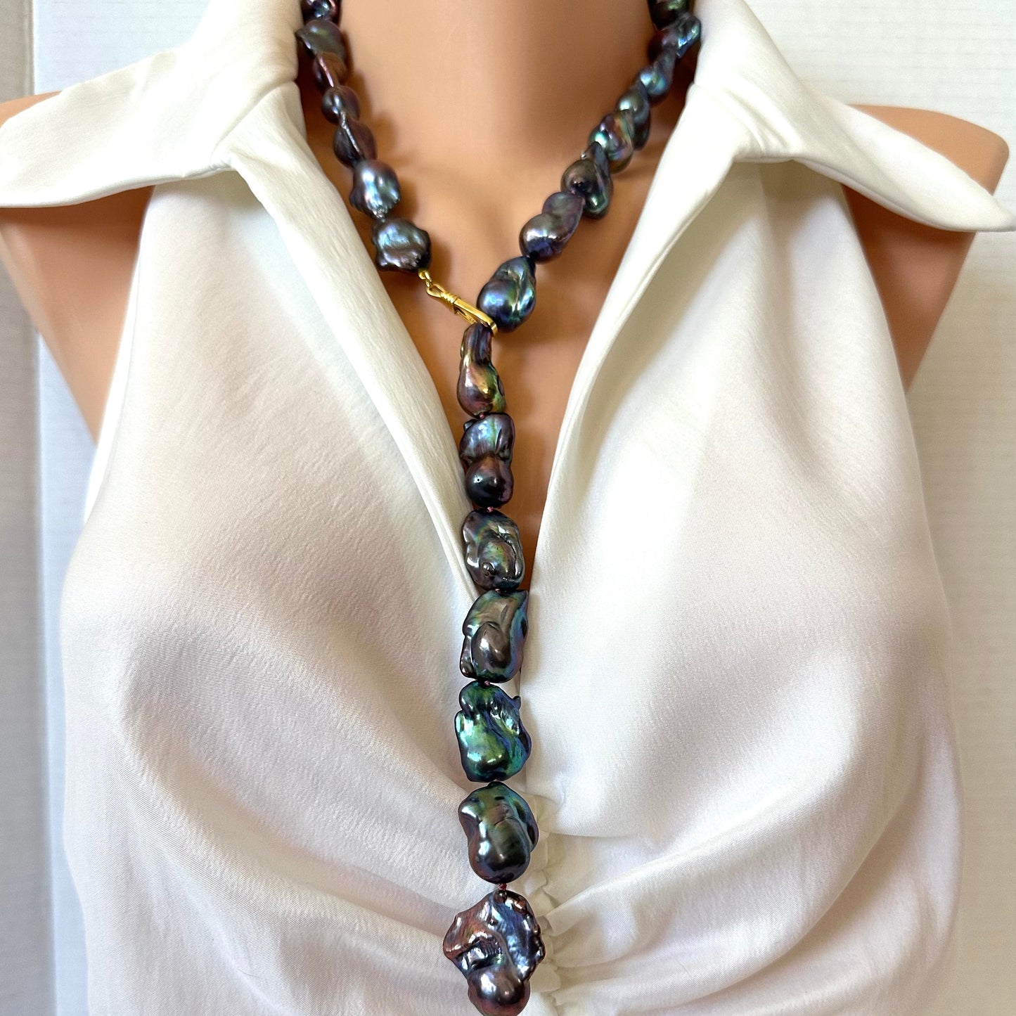 A close-up of a baroque pearl necklace with iridescent green, blue, and purple tones, styled on a mannequin wearing a cream satin top.