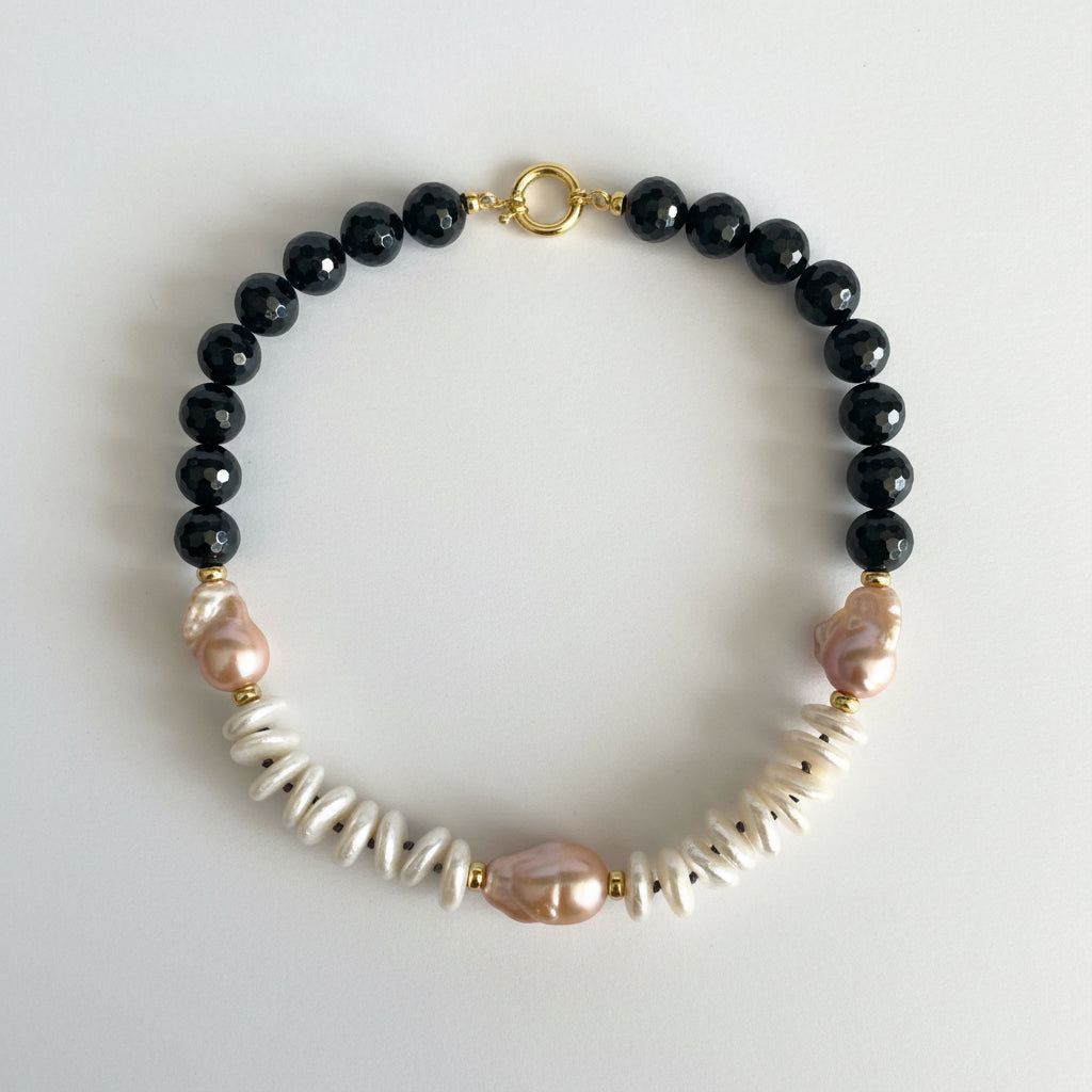 A beaded necklace featuring faceted black beads, irregular white shell beads, and natural pink baroque pearls, accented with small gold spacers and a gold clasp, displayed on a clean white background.