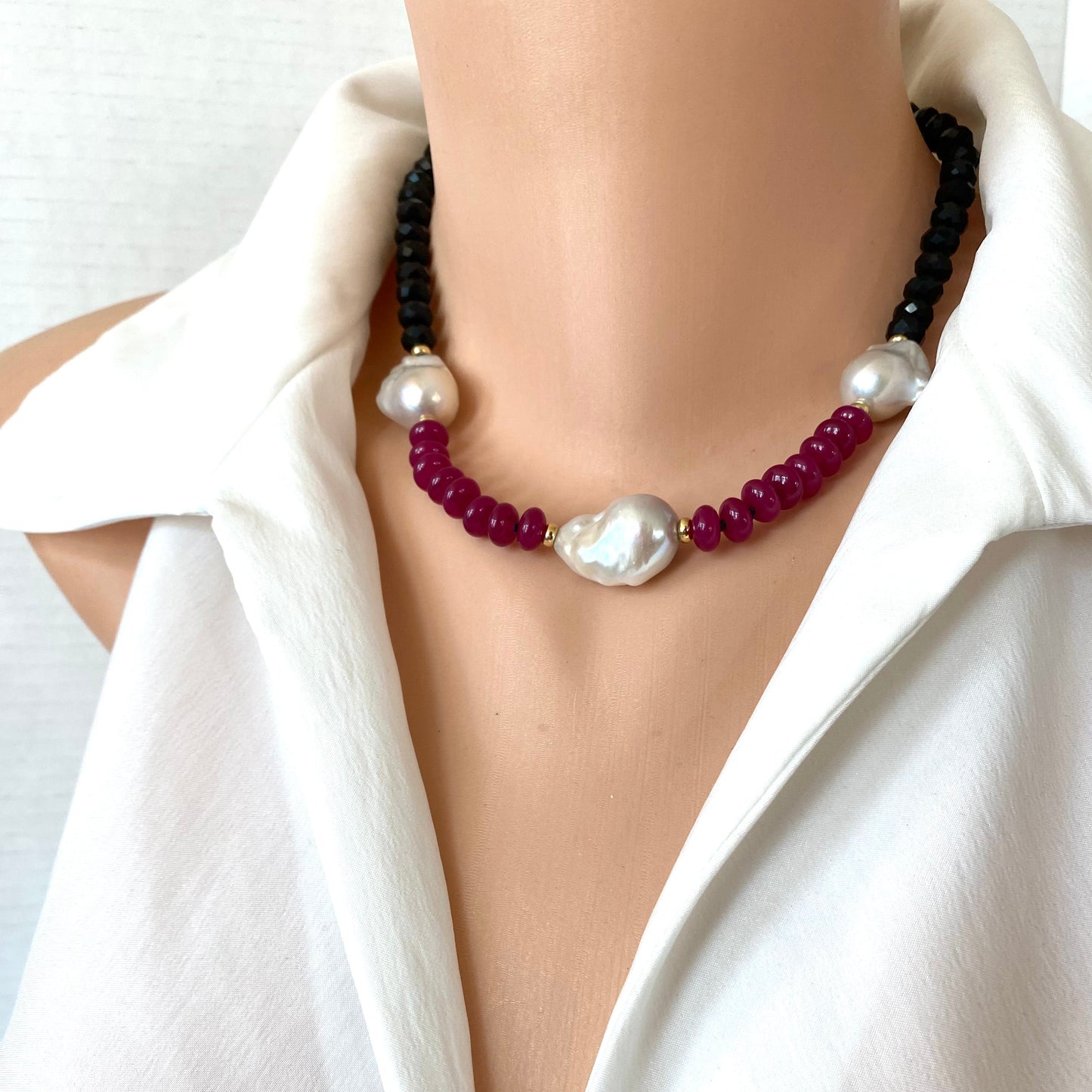 A close-up of a beaded necklace displayed on a mannequin torso wearing a white blouse, dark black beads with deep red beads, accented by white pearls.