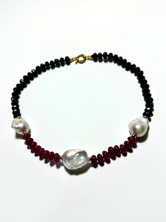A beaded necklace featuring black faceted beads, deep red round beads, large irregular white baroque pearls, and a gold clasp, displayed on a clean white background.