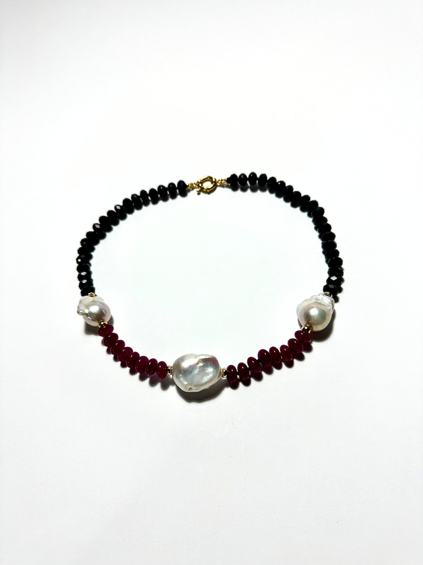 A beaded necklace featuring black faceted beads, deep red round beads, and three large white baroque pearls, with a gold clasp, displayed on a clean white background.