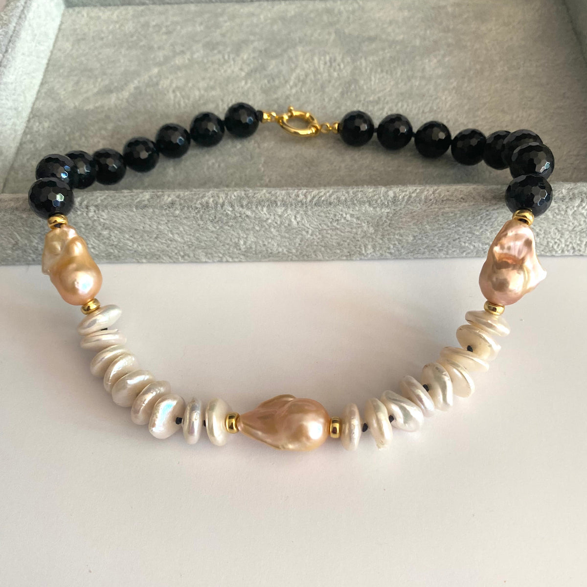 A beaded necklace featuring black faceted beads, white baroque pearls, and pale pink baroque pearls, separated by small gold spacer beads, displayed on a gray velvet jewelry box.