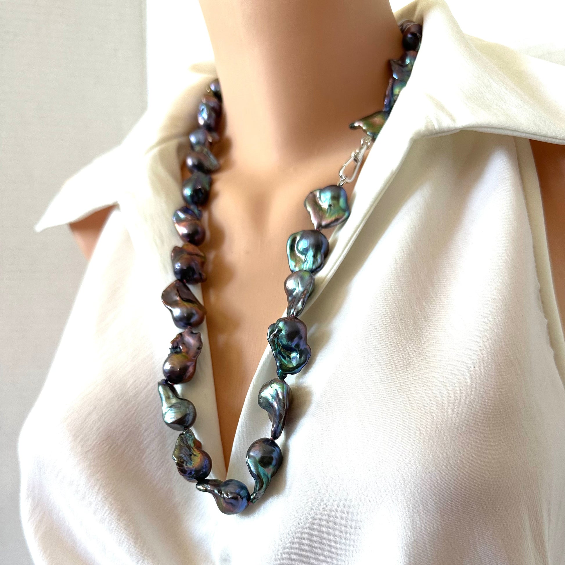 A close-up of a necklace displayed on a mannequin torso wearing a white blouse. The necklace is made of irregular, polished baroque-style shell beads with a dark iridescent finish that shifts between blue, green, and purple.