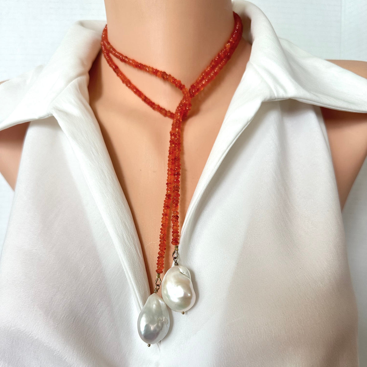 A close-up of a beaded lariat-style necklace with two large baroque pearls, worn on a mannequin torso wearing a white blouse.