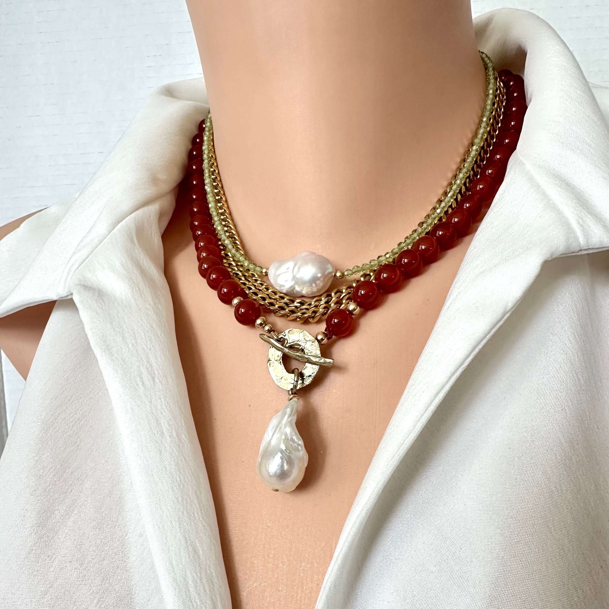 A close-up of a multi-strand necklace worn on a mannequin torso dressed in a white shirt. The necklace features red beads, gold-tone chains, a white pearl, and a gold toggle clasp with a teardrop pearl pendant.