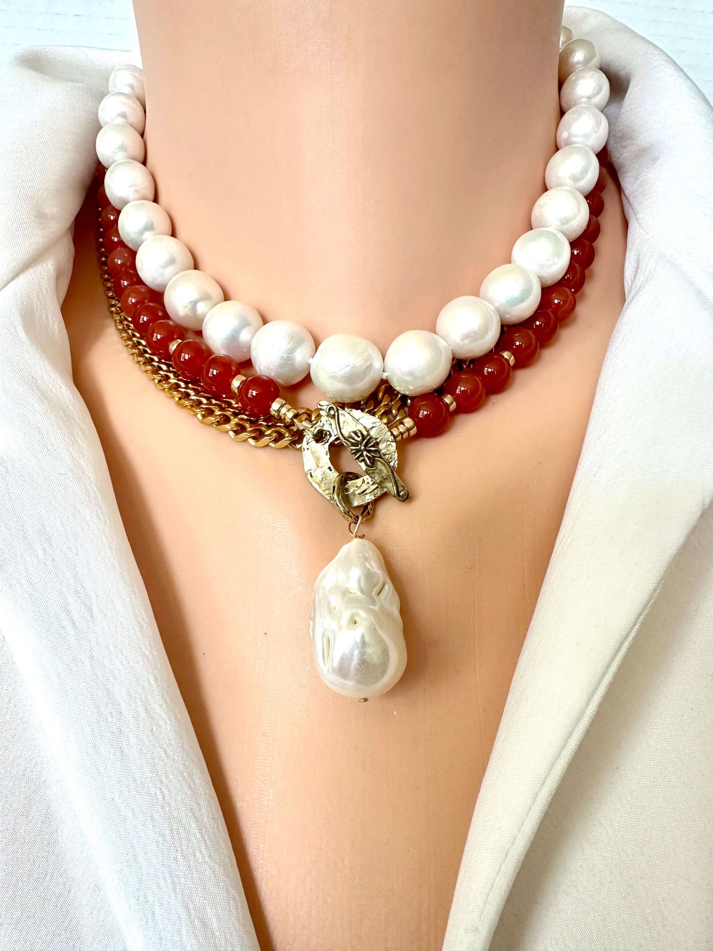 A close-up of a multi-strand necklace displayed on a mannequin bust. The necklace features a top strand of large white baroque pearls, a middle strand of glossy red beads, a gold-tone chain, and a central antique gold clasp holding a large white teardrop pearl pendant.