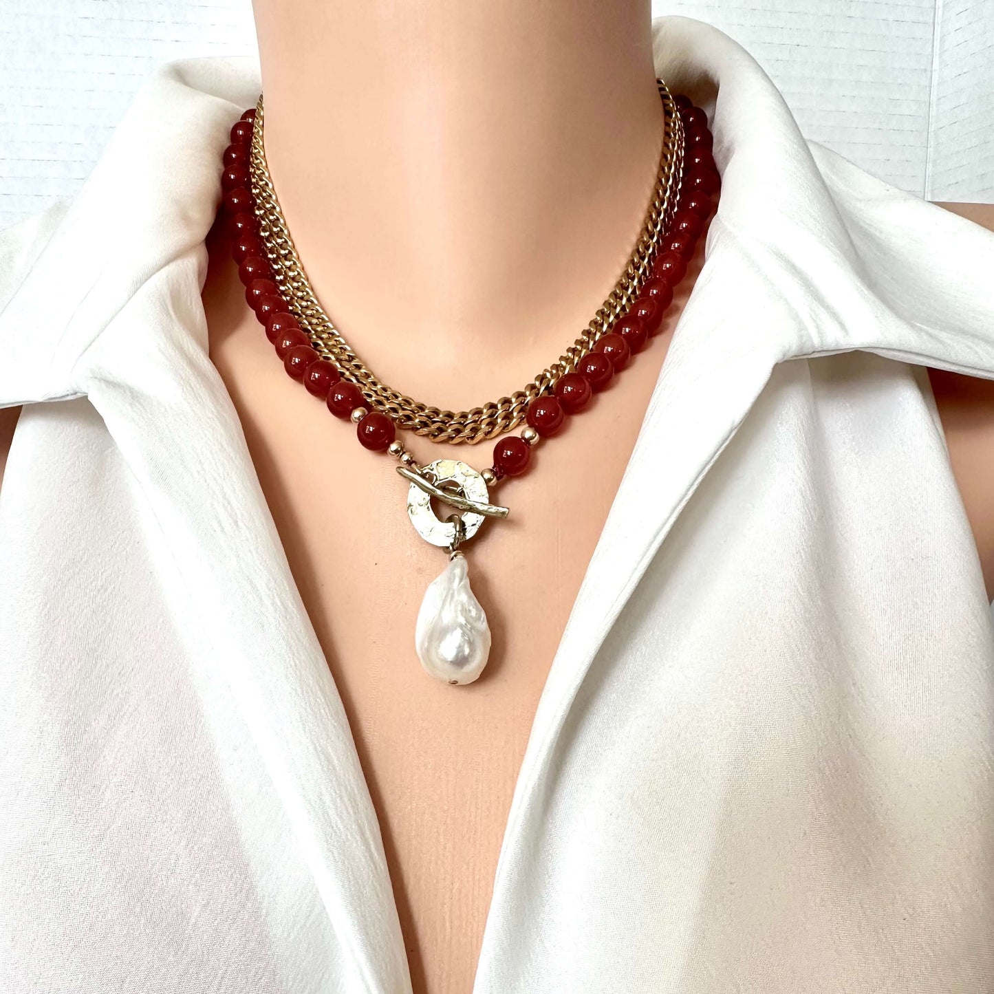 A close-up of a double-strand necklace worn on a mannequin torso. The necklace features a top layer of gold-tone chain and a bottom layer of dark red beads, with a silver-tone toggle clasp holding a white baroque pearl pendant.