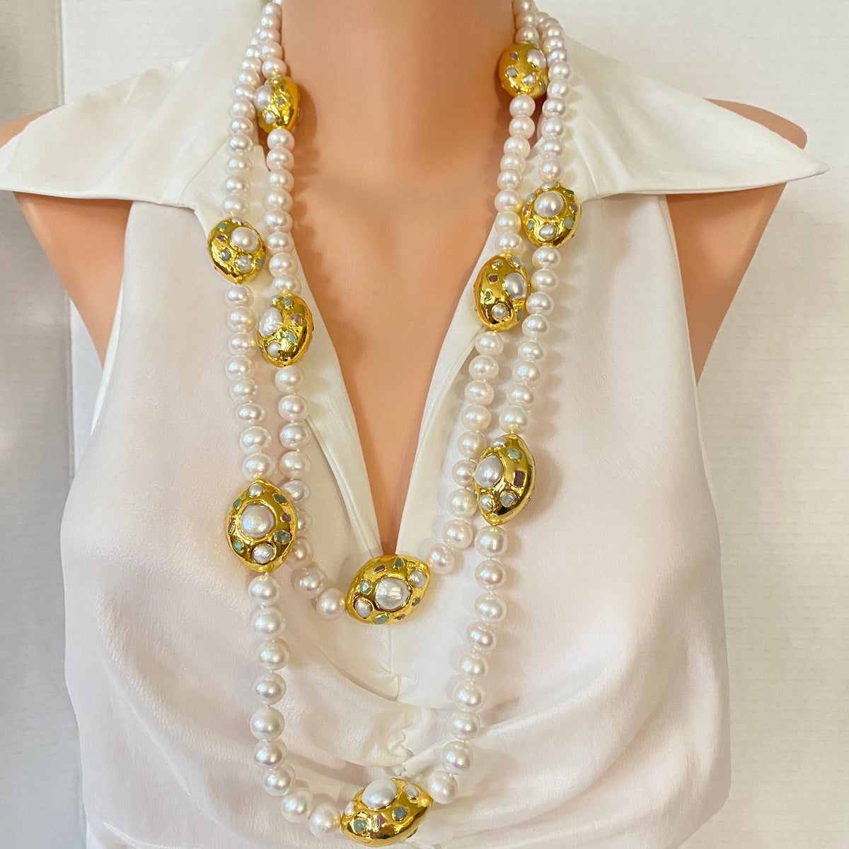 A double-strand pearl necklace with ornate gold-tone oval medallions, displayed on a cream-colored satin dress form.