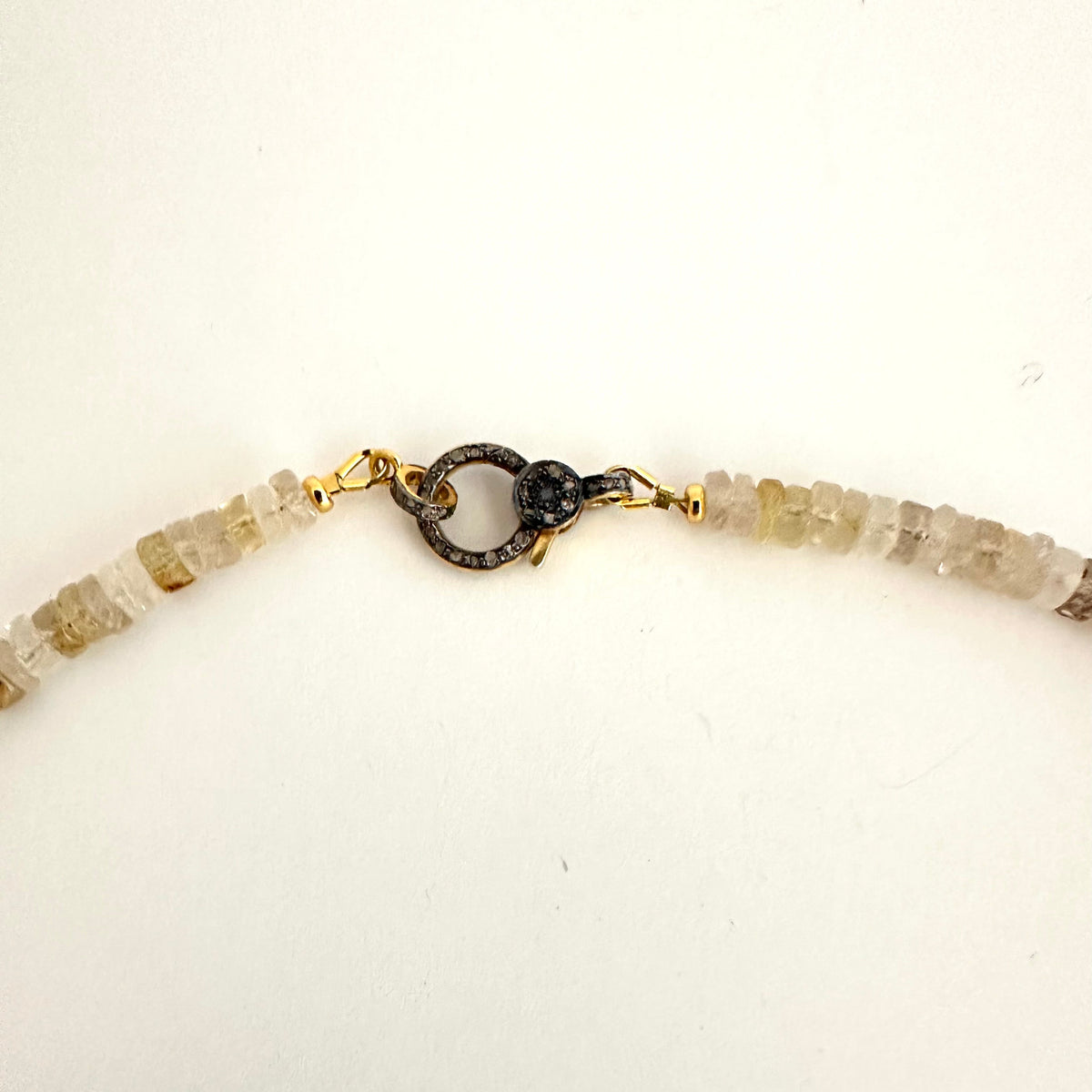 A close-up of a beaded necklace with translucent pale cream and light brown beads, featuring a gold-toned lobster clasp with a dark textured circular accent, laid on a plain off-white background.