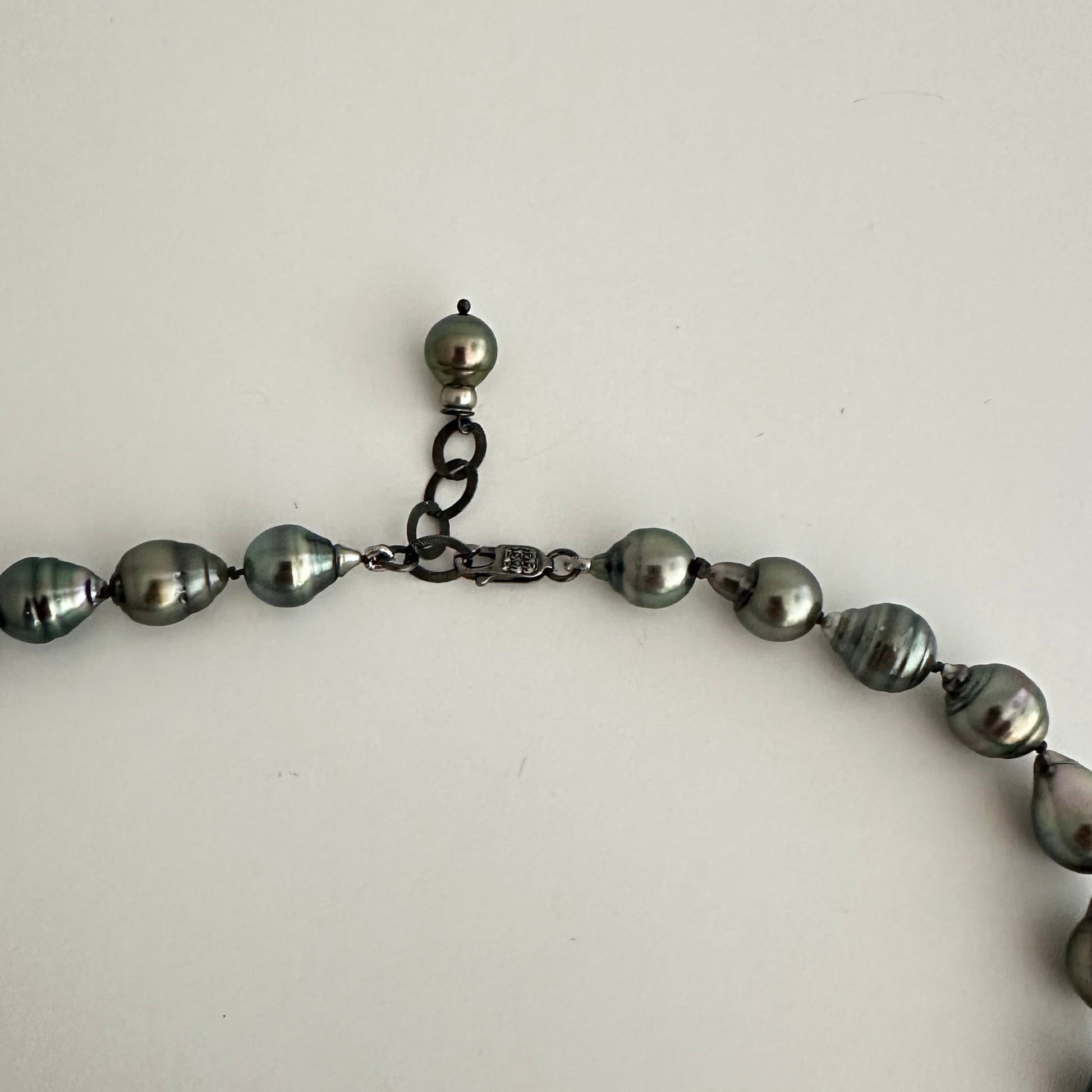 A close-up of a pearl necklace with dark gray, baroque-shaped pearls and a dark metal clasp, laid against a plain off-white background.