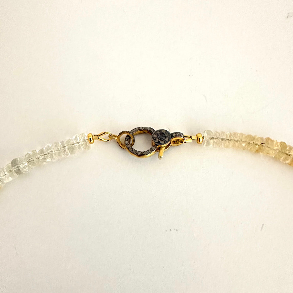 A close-up of a beaded necklace with clear faceted beads and a gold-toned clasp on a plain off-white background.