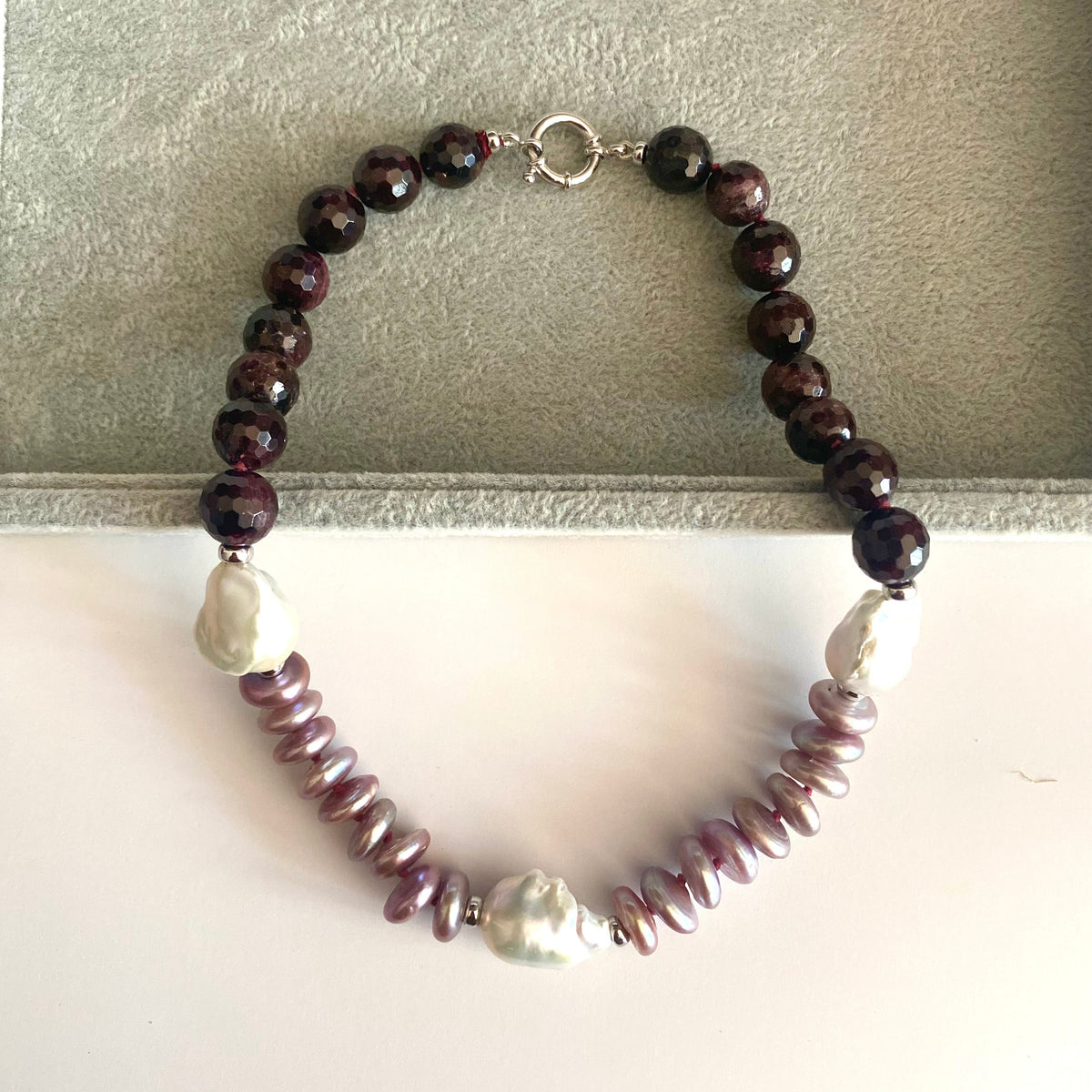 A beaded necklace with dark reddish-brown faceted beads, light purple baroque pearls, and a strand of small lavender beads, displayed on a gray jewelry box.