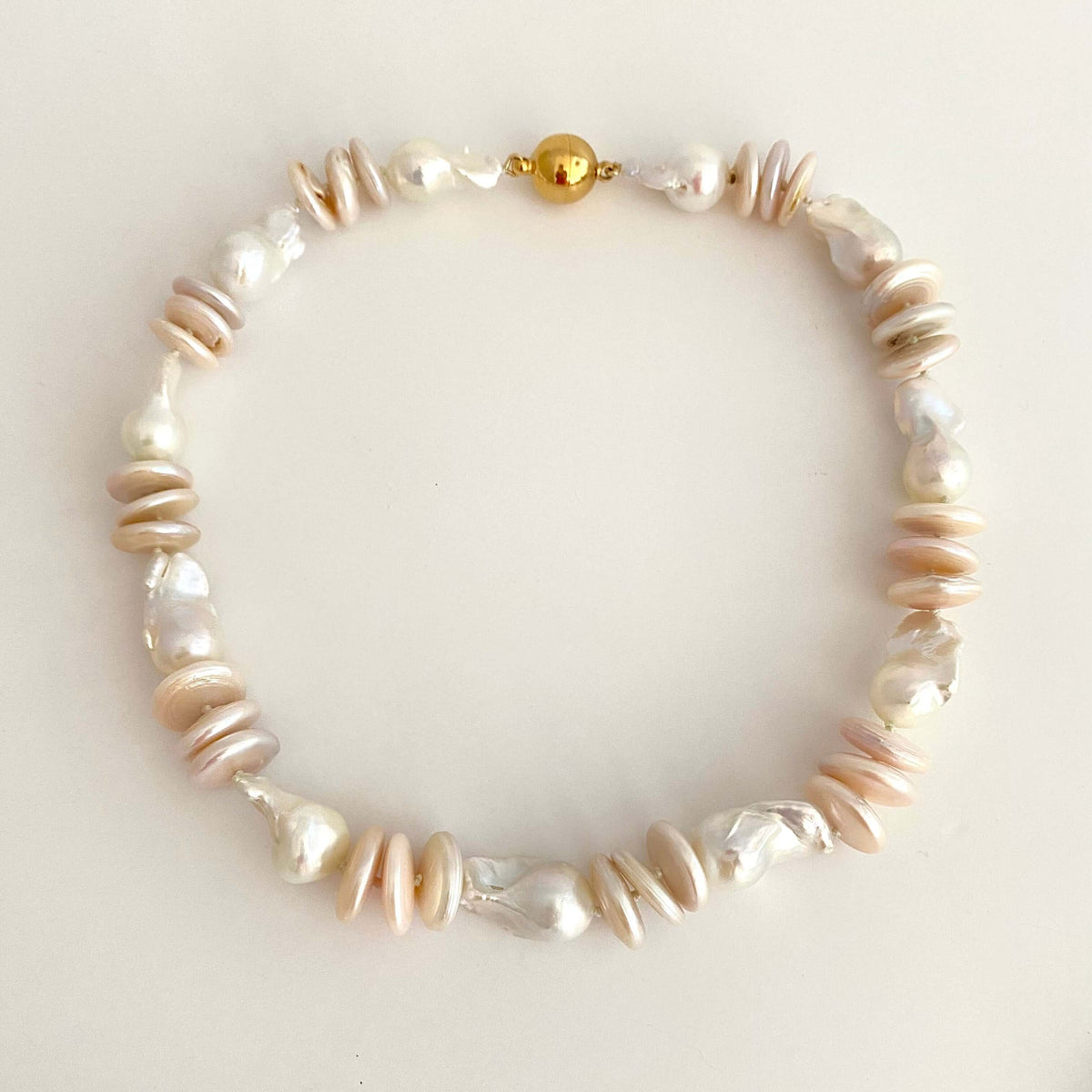 A beaded necklace featuring a mix of natural baroque pearls and flat, disc-shaped shell beads in cream, beige, and white, accented by a small gold clasp, displayed on a light cream background.