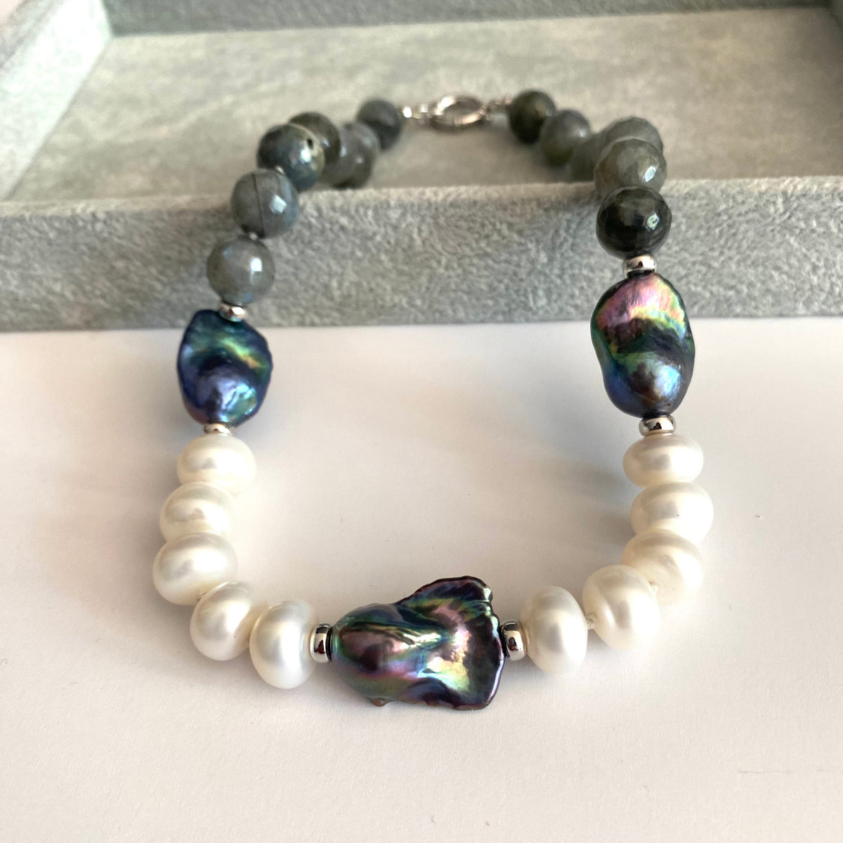 A beaded necklace featuring white baroque pearls, dark gray beads, and three large iridescent green-blue baroque shell beads, displayed on a light surface.