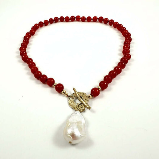 A beaded necklace featuring deep red round beads, a gold-toned toggle clasp, and a large, irregular white baroque pearl pendant, displayed on a clean white background.