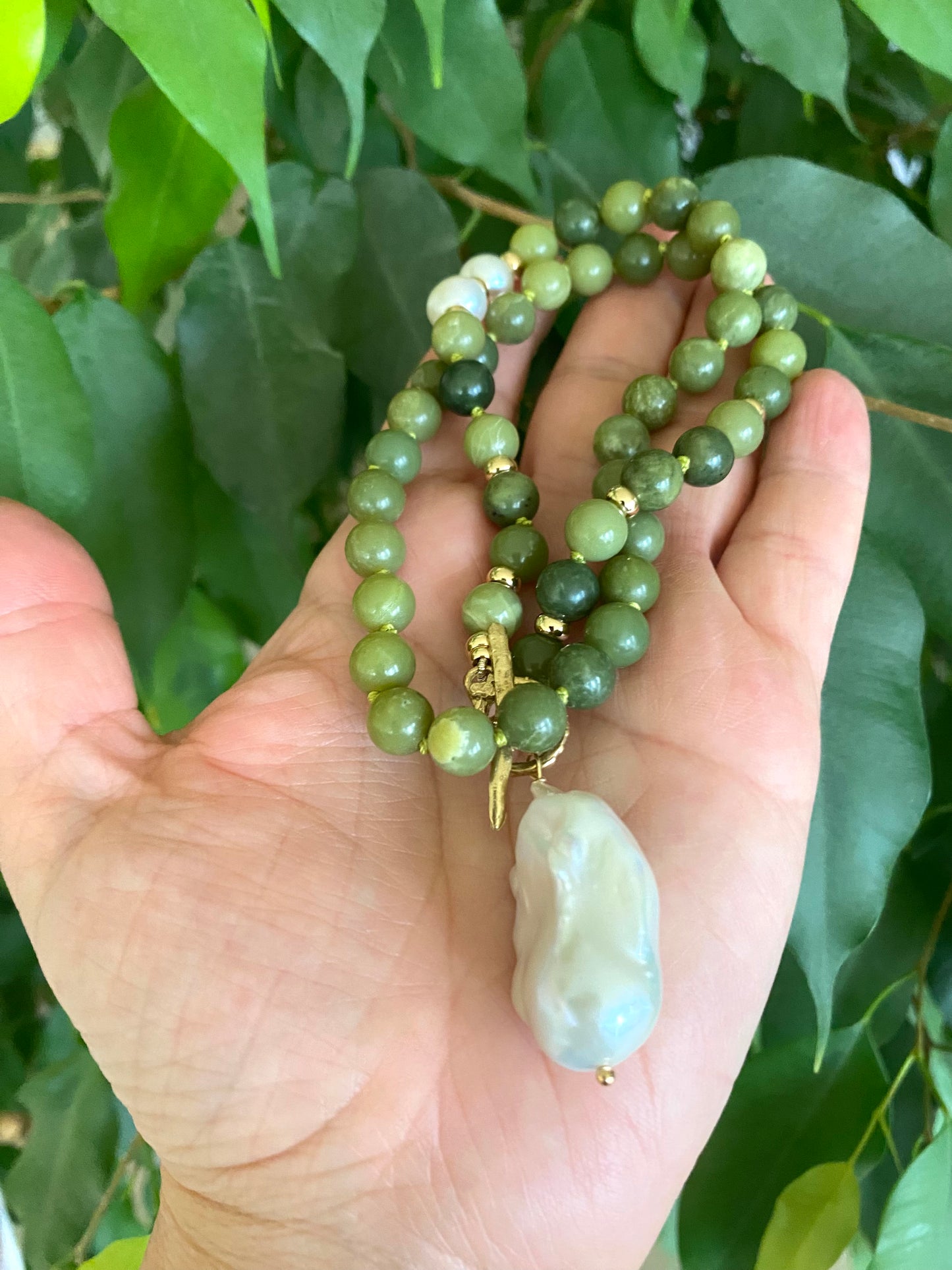 Green beaded necklace with a large white pearl held in a hand against a green leafy background