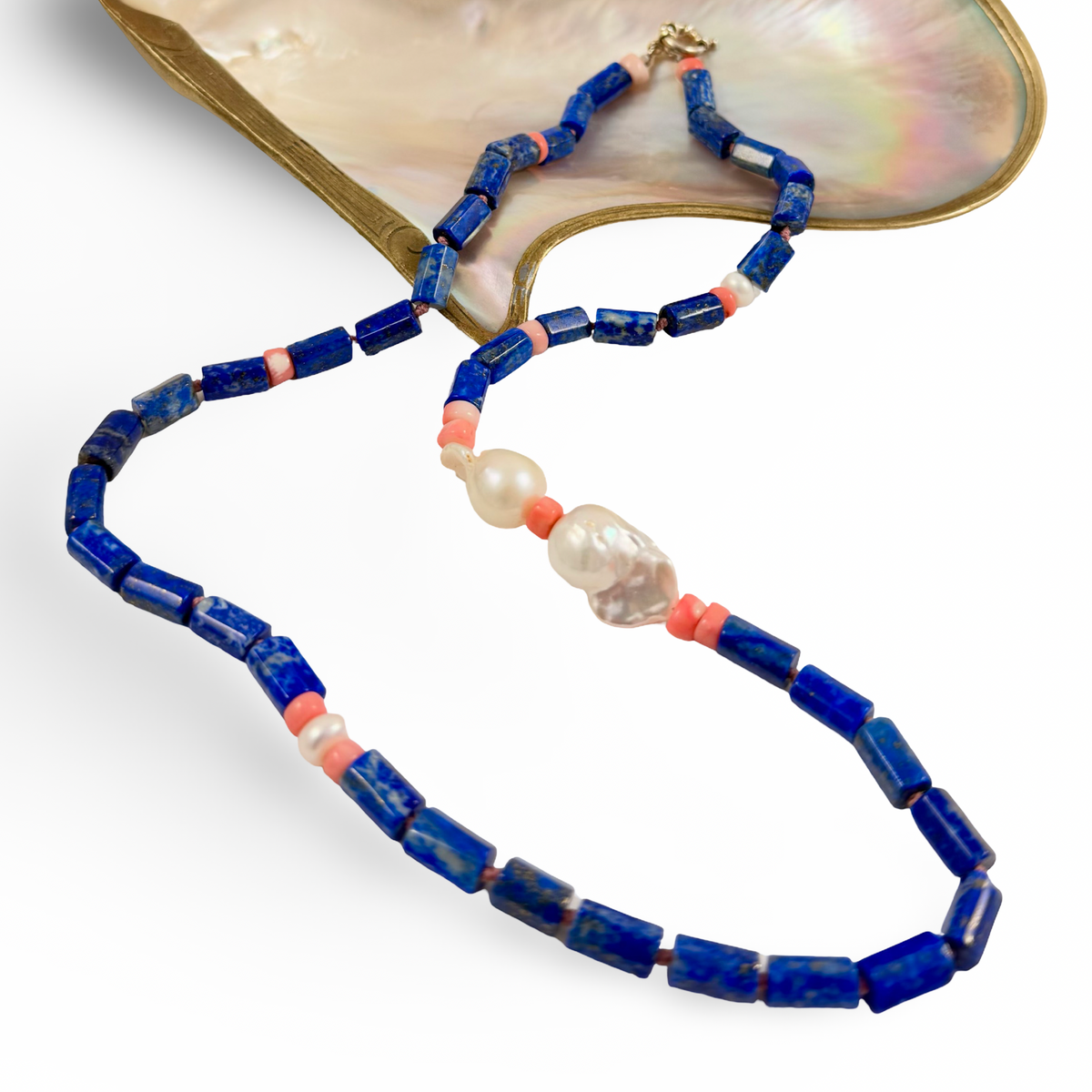 A beaded necklace with dark blue lapis lazuli-style rectangular beads, coral orange spacer beads, white pearl-like beads, and a clear faceted bead, displayed on a white background with a mother-of-pearl shell prop.