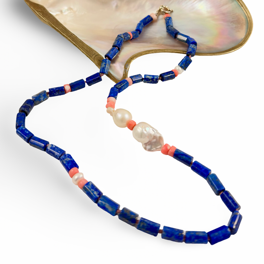 A beaded necklace with dark blue lapis lazuli-style rectangular beads, coral orange spacer beads, white pearl-like beads, and a clear faceted bead, displayed on a white background with a mother-of-pearl shell prop.