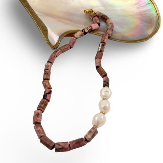A beaded necklace featuring chunky terracotta-pink stone beads with black marbling, accented by three smooth white baroque pearls and a small gold clasp, displayed on a white background with a mother-of-pearl shell prop.