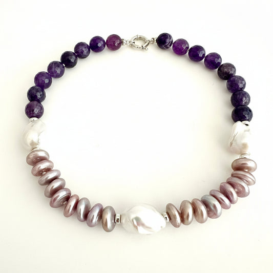 A beaded necklace featuring dark purple amethyst beads on one side, large white baroque pearls on the other, and iridescent pinkish-brown baroque pearls in the center, displayed on a plain off-white background.