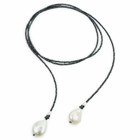 A long-strand beaded necklace with dark gray beads and two large, irregular white baroque pearls at the ends, displayed on a clean white background.