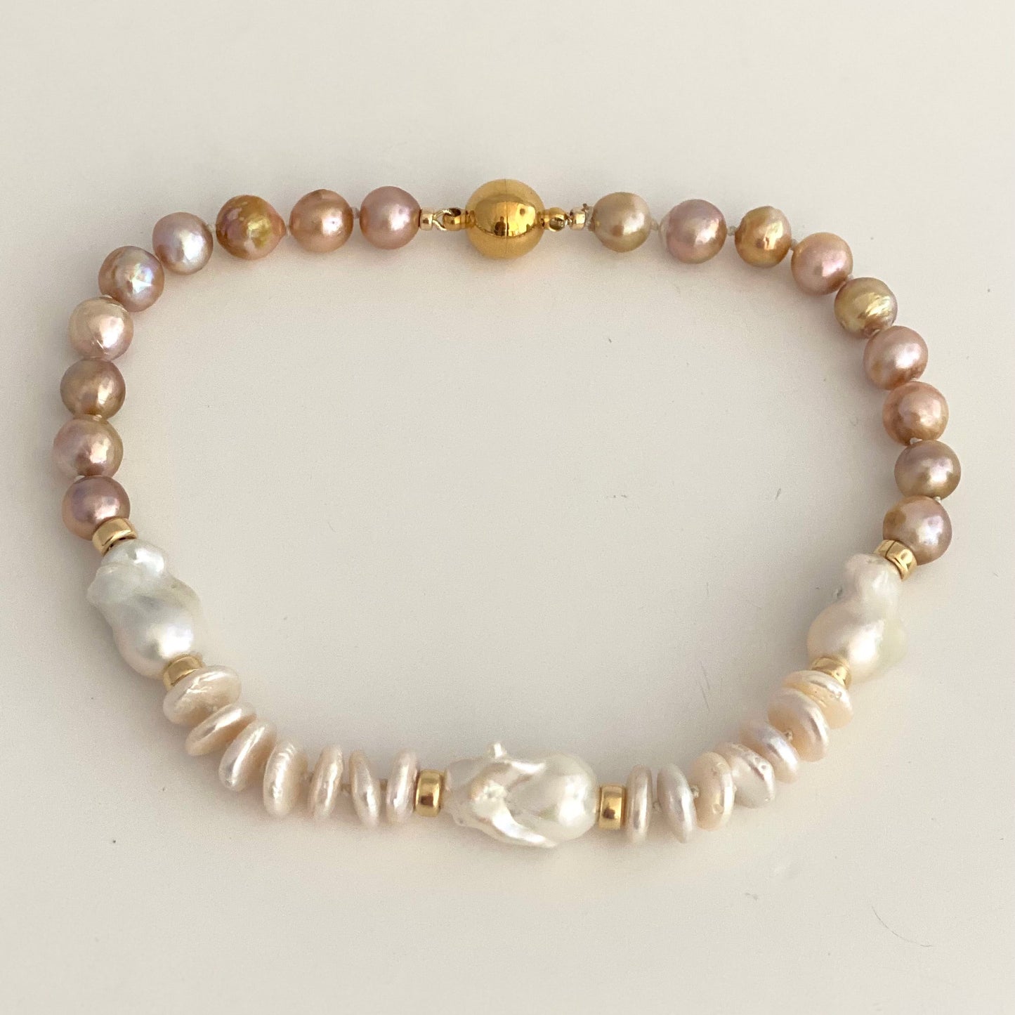 A beaded pearl bracelet featuring a mix of iridescent pinkish-gold, white baroque, and textured white pearls, accented with small gold spacer beads and a central gold bead clasp, displayed on a light cream background.
