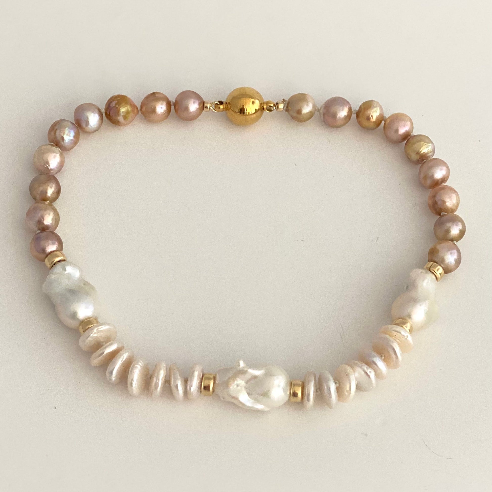 A beaded pearl bracelet featuring a mix of iridescent pinkish-gold, white baroque, and textured white pearls, accented with small gold spacer beads and a central gold bead clasp, displayed on a light cream background.