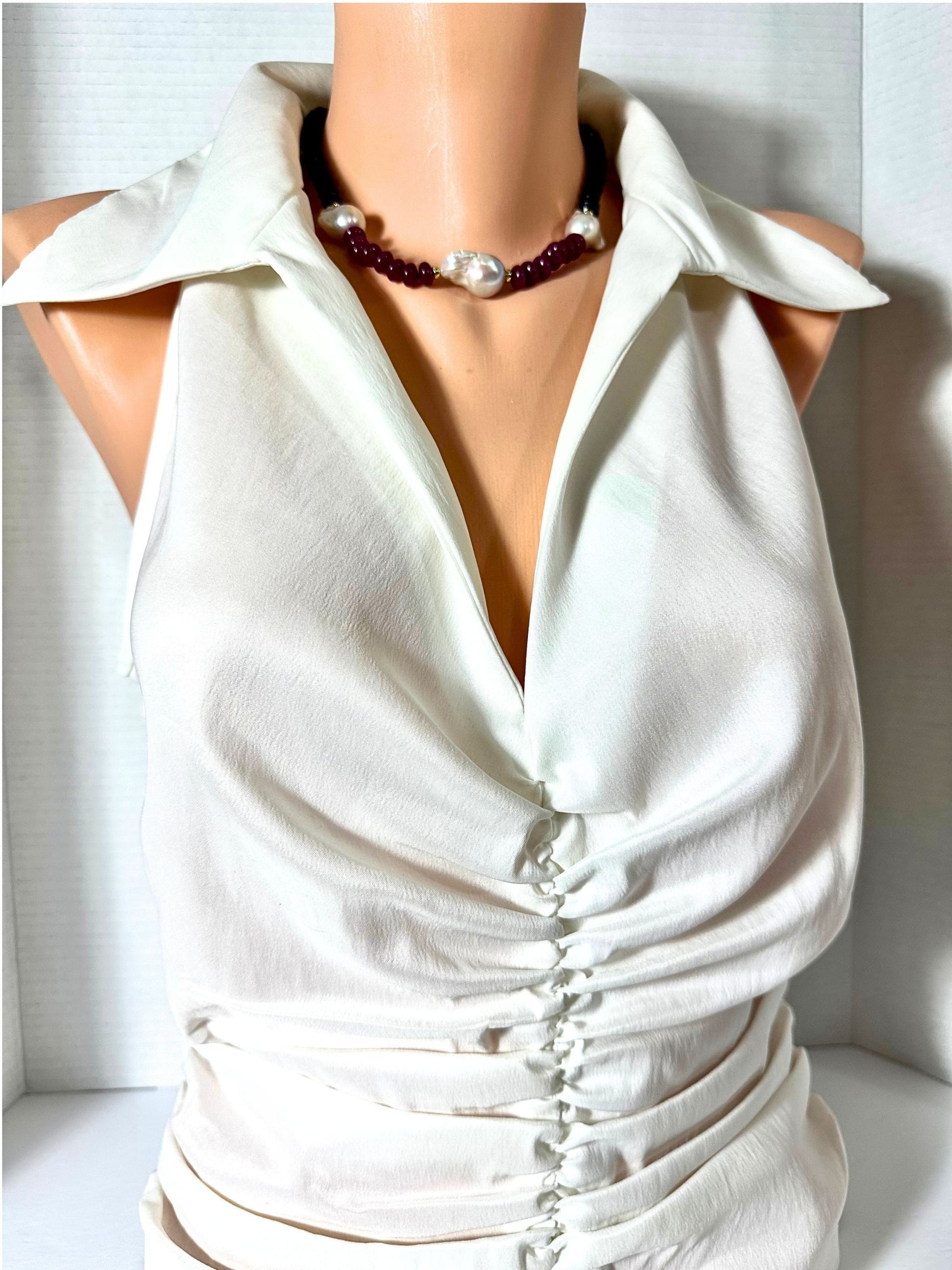 An off-white sleeveless blouse with a deep V-neckline, shoulder cutouts, and a front button closure, displayed on a mannequin torso with a beaded necklace.