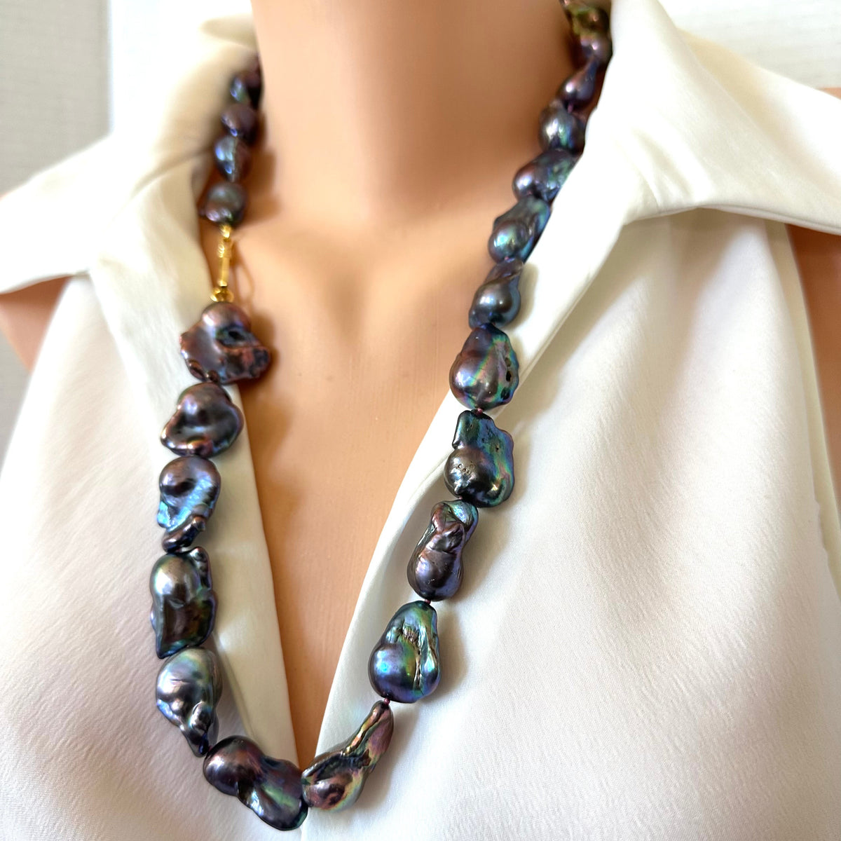 A close-up of a baroque pearl necklace with dark iridescent beads and a gold clasp, displayed on a mannequin torso wearing a white blouse.