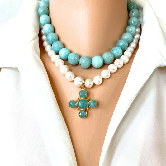 A close-up of a double-strand necklace worn on a mannequin or model's neck, featuring a top layer of light blue round beads and a bottom layer of white faux pearls, with a small gold cross pendant set with light blue stones at the center. The necklace is styled with a white collared shirt.