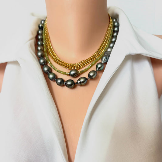A close-up of a model's neck and upper chest wearing a layered necklace set over a white collared blouse. The jewelry includes dark gray pearl strands, gold chain layers, and a green beaded accent chain.