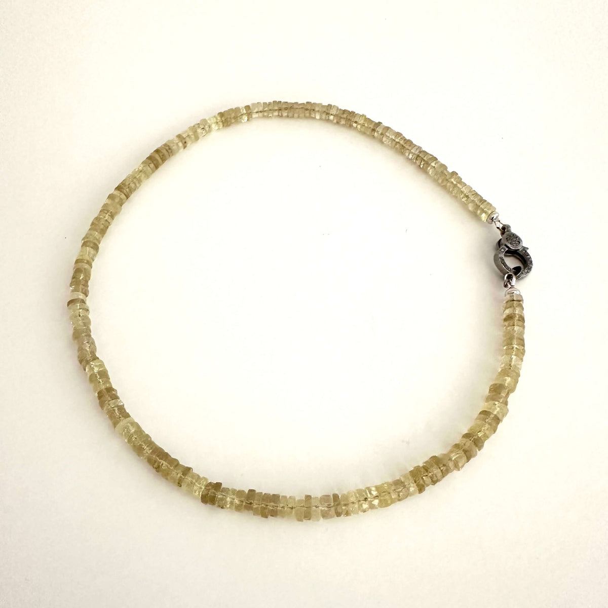 A beaded necklace made of small, faceted translucent pale yellow beads, arranged in a circle on a plain white background. It features a dark metal lobster clasp at the end.
