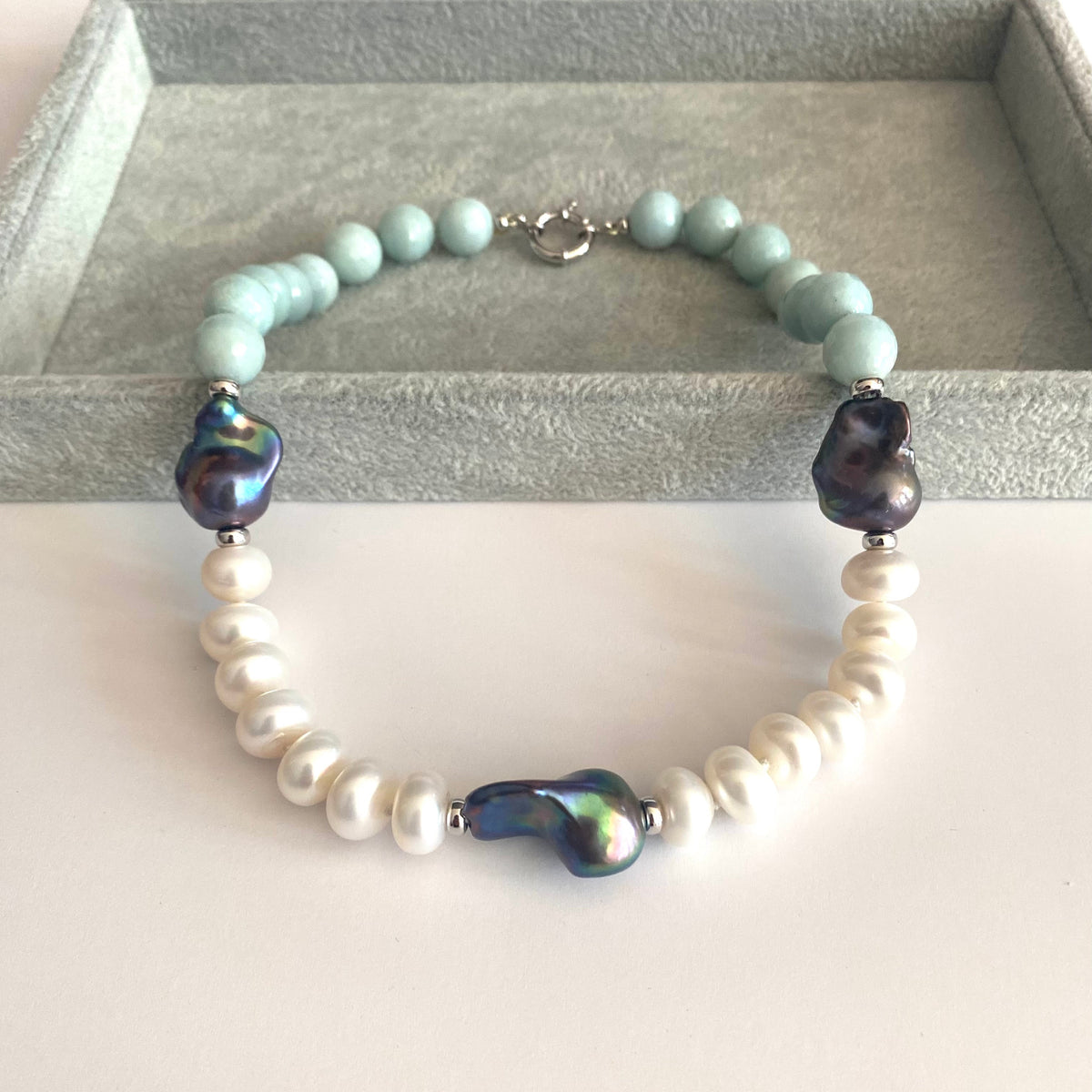 A beaded bracelet displayed on a gray jewelry box. The bracelet features light blue-green round beads, white baroque pearls, and three dark iridescent beads, with a silver clasp.
