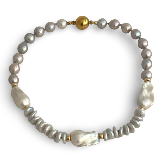 hand knotted necklace featuring a strand of 10mm gray pearls, a few larger white baroque pearls, coin gray beads, and gold spacer accents, displayed on a light beige background.