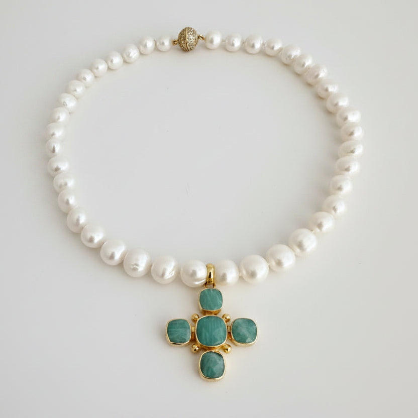 A pearl beaded necklace with a gold cross pendant featuring green cabochon stones, displayed on a light gray background.