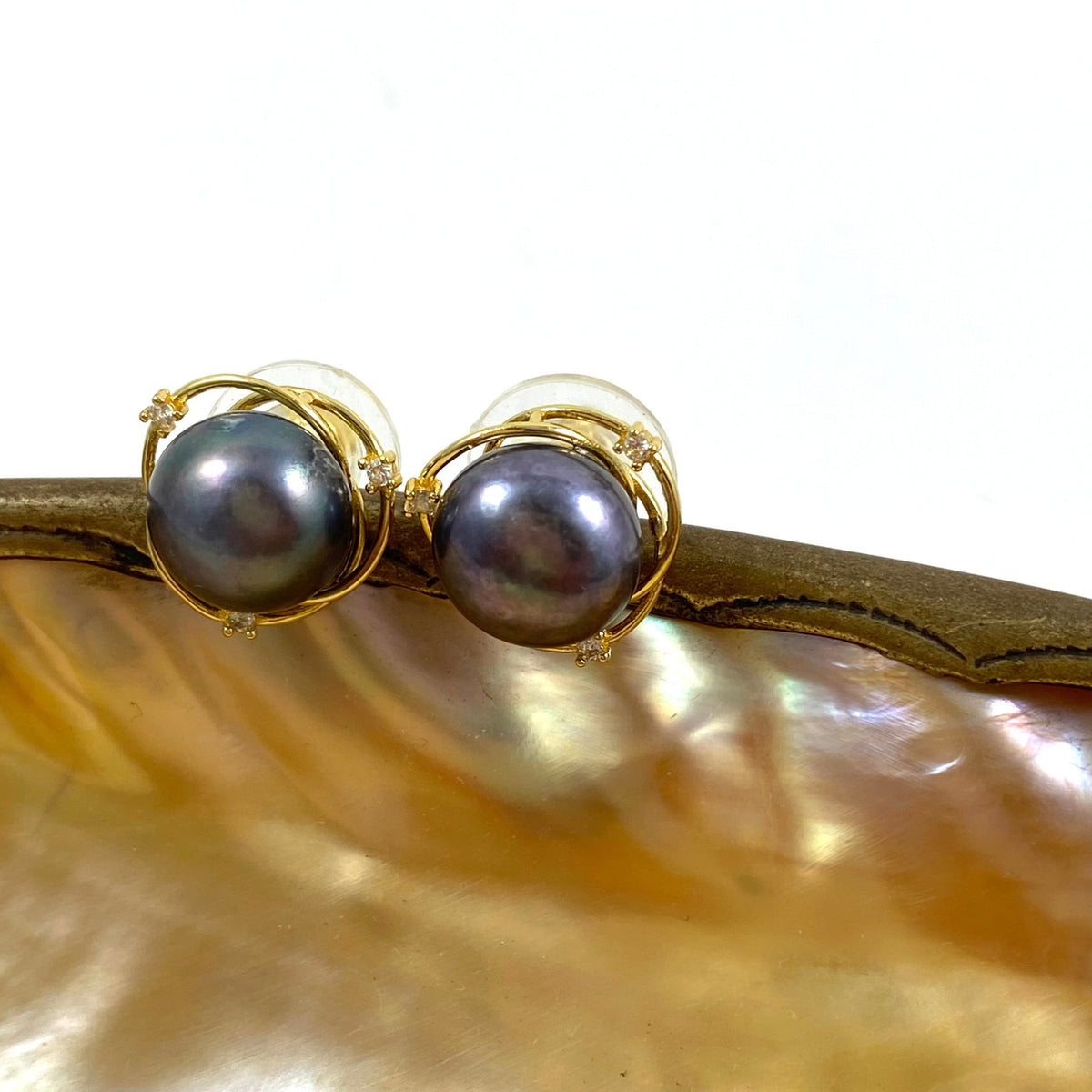 A pair of gold stud earrings with round dark gray pearls, resting on a curved piece of polished mother-of-pearl shell against a white background.