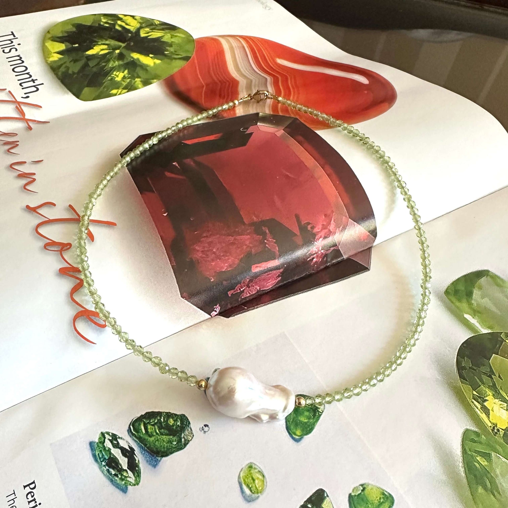 A delicate beaded bracelet with a baroque pearl and green gemstone accents, displayed on an open book surrounded by gemstone samples.